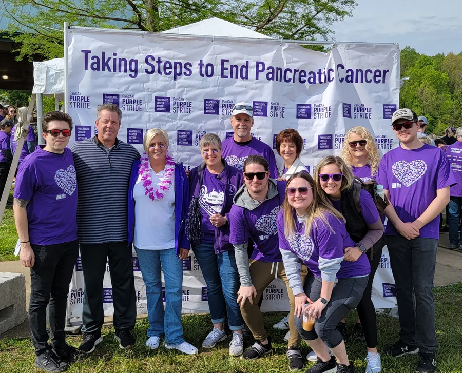Connie&rsquo;s Crusaders supporters in purple shirts beneath a PanCAN PurpleStride banner