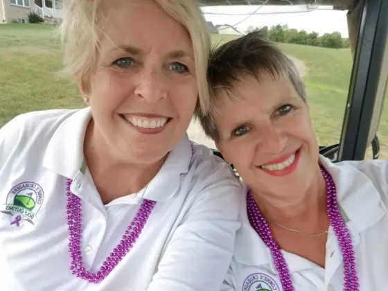 Two smiling women in white golf polos and purple beads in a golf cart at the golf outing