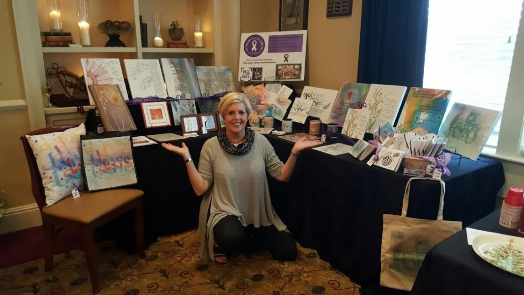 A display of Connie&rsquo;s Creations on black-draped tables: canvases, framed pieces, note cards, a pillow and a tote bag printed with her artwork, with a smiling woman kneeling in front